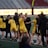 A group of athletes wearing yellow sports uniforms are standing on a sports field. Some are holding lacrosse sticks, and they appear to be interacting casually. Several people are dressed in black coats. In the background, there is a bright sign with text and parts of a stadium are visible.