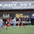 Players engage in a competitive soccer game on a field while others are seated on the bench watching attentively. The action is taking place in front of a wooden structure displaying multiple advertisements, including orthodontics, lacrosse, and field hockey.
