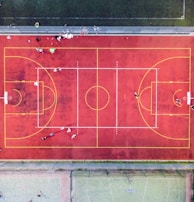 An aerial view of a sports court with a vibrant red surface and yellow lines marking different sections for basketball and soccer. Several people are present on the court, some engaged in activities, while others are standing along the perimeter. The court is enclosed by a metal fence and surrounded by grass on all sides.