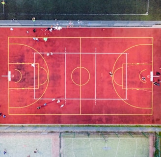 An aerial view of a sports court with a vibrant red surface and yellow lines marking different sections for basketball and soccer. Several people are present on the court, some engaged in activities, while others are standing along the perimeter. The court is enclosed by a metal fence and surrounded by grass on all sides.